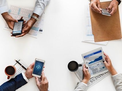 Four people using mobile phones on a desk with financial forms, papers and newspapers lying around