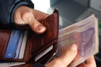 A person holding a brown leather wallet and banknotes in front of an ATM machine