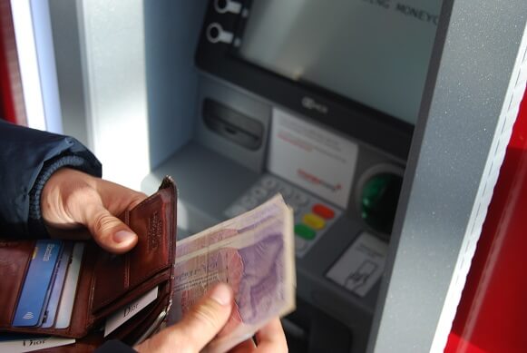 A person holding a brown leather wallet and banknotes in front of an ATM machine