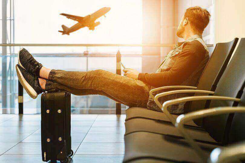 A man sitting on gang chair at the airport with feet propped up on his luggage, looking at airplane taking off