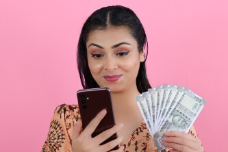 An image of a happy woman with a mobile phone in one hand and a fan of Indian currency notes in the other hand