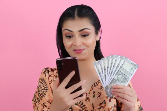 An image of a happy woman with a mobile phone in one hand and a fan of Indian currency notes in the other hand
