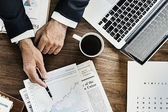 A man presenting investment data on sheets of paper alongside a cup of coffee, a laptop and other papers lying on a desk