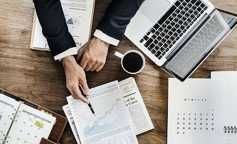 A man presenting investment data on sheets of paper alongside a cup of coffee, a laptop and other papers lying on a desk